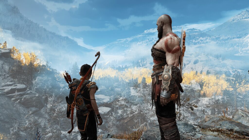 Father and son from the God of War video game series stand at the top of a mountain and look down on a snowy landscape.