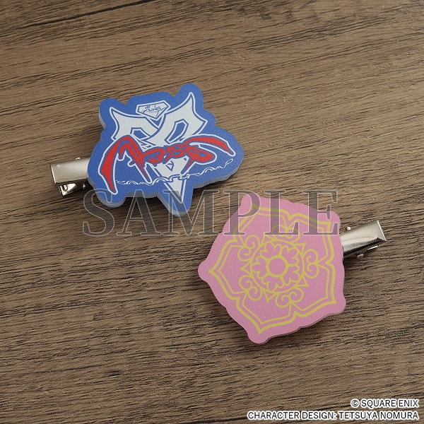Two flat acrylic hair clips on a wooden surface, one with the Zanarkand Abes Blitzball team logo, the other with Yuna's hibiscus design.